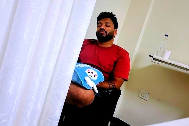 A father sitting on a chair in a hospital, holding a blue blanket with a character design, appearing relaxed with eyes closed.